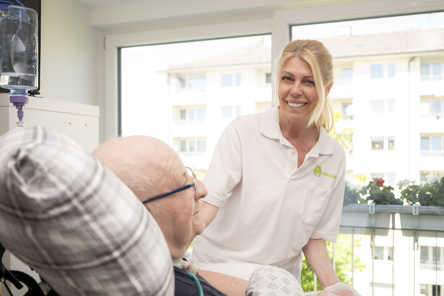 Eine lächelnde VIVICARE Pflegekraft beugt sich über einen älteren, ebenfalls lächelnden Patienten im Bett und betreut ihn.